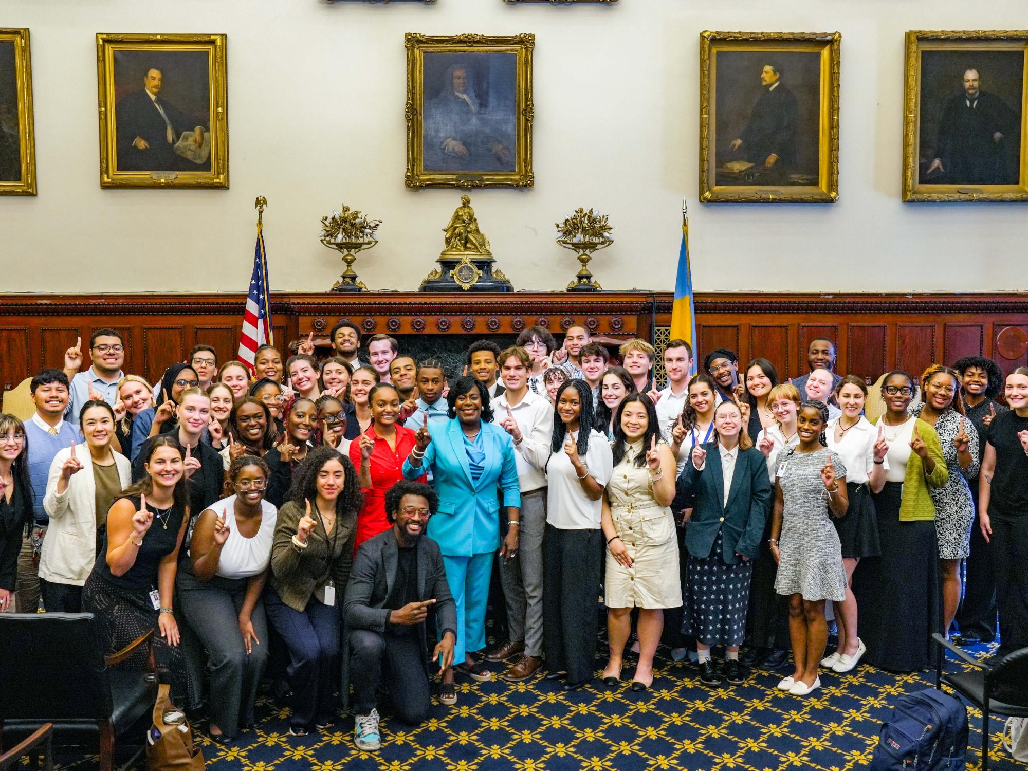 A large group of diverse students and professionals pose together in a formal room, smiling and holding up one finger. They stand in front of a fireplace with framed portraits on the wall, and American and Ukrainian flags displayed behind them.