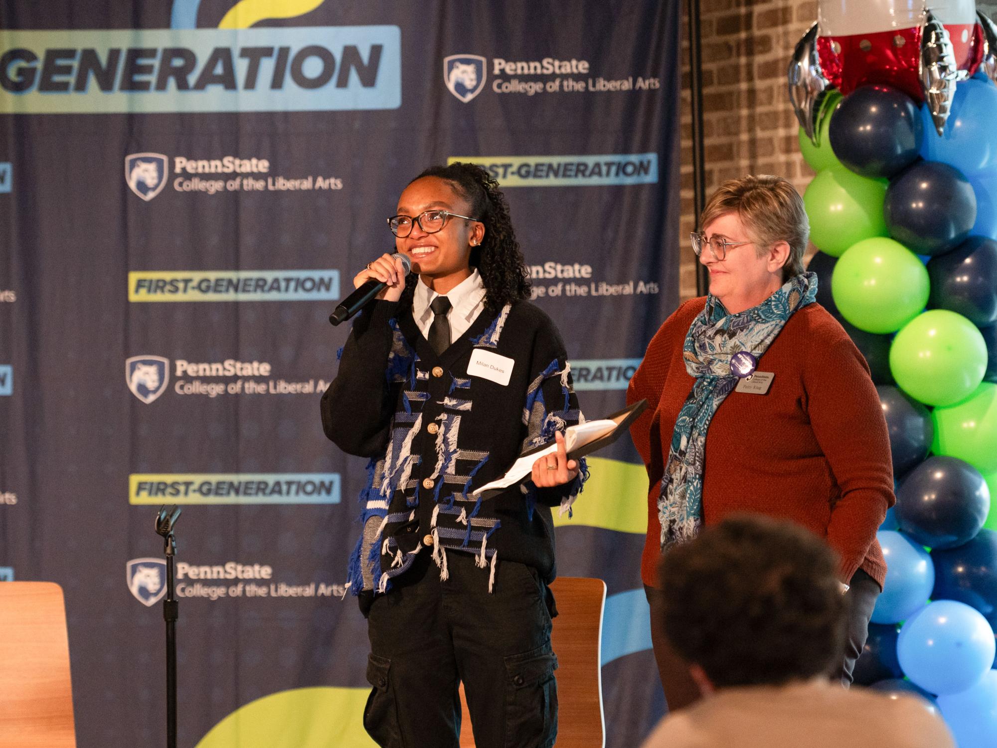 Milan Dukes stands on stage holding a microphone and speaking at a Penn State Liberal Arts First-Generation event, with a banner behind her and Patty Klug standing to her right.
