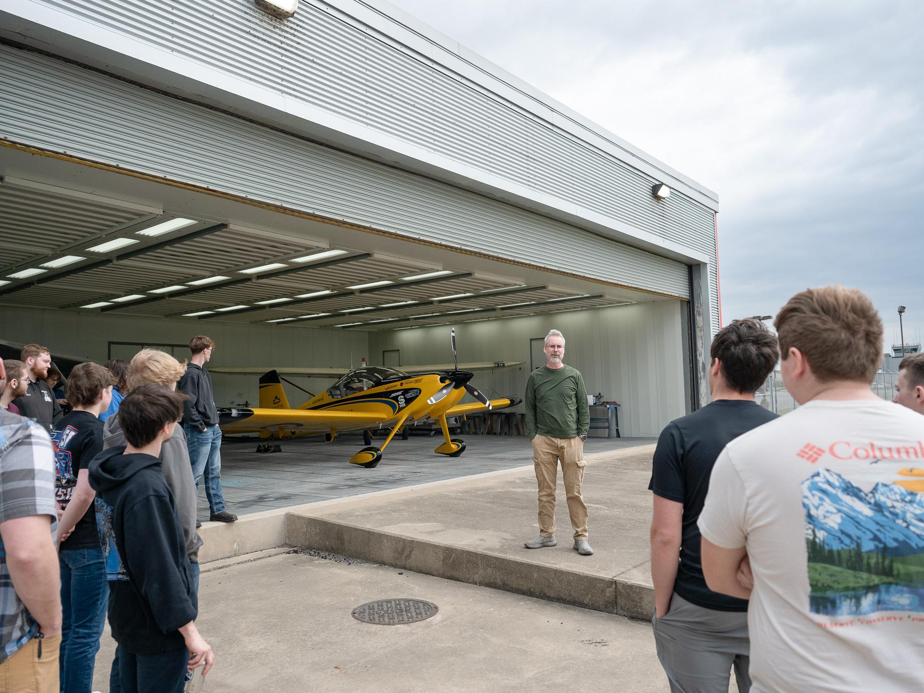 A group of aviation students listen to a pilot in the entrance to a hangar