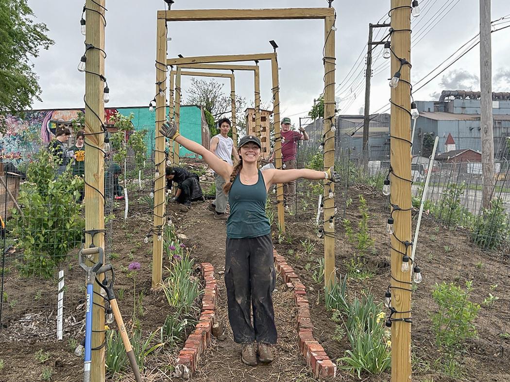 Person standing with arms outstretched beneath a series of wooden garden archways wrapped with string lights, with other volunteers gardening along a narrow brick path behind them.