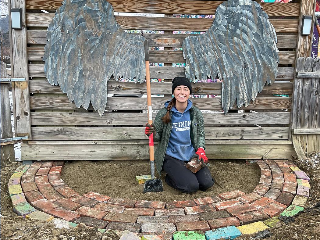 Person kneeling in front of a wooden fence with large metal wing sculptures, holding a shovel beside a small circular brick seating area in a garden.