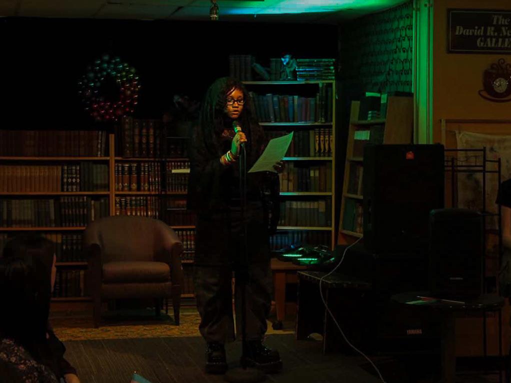 Zach Hamer-Lang, wearing a black jacket and black pants, standing in a dark room holding a microphone.