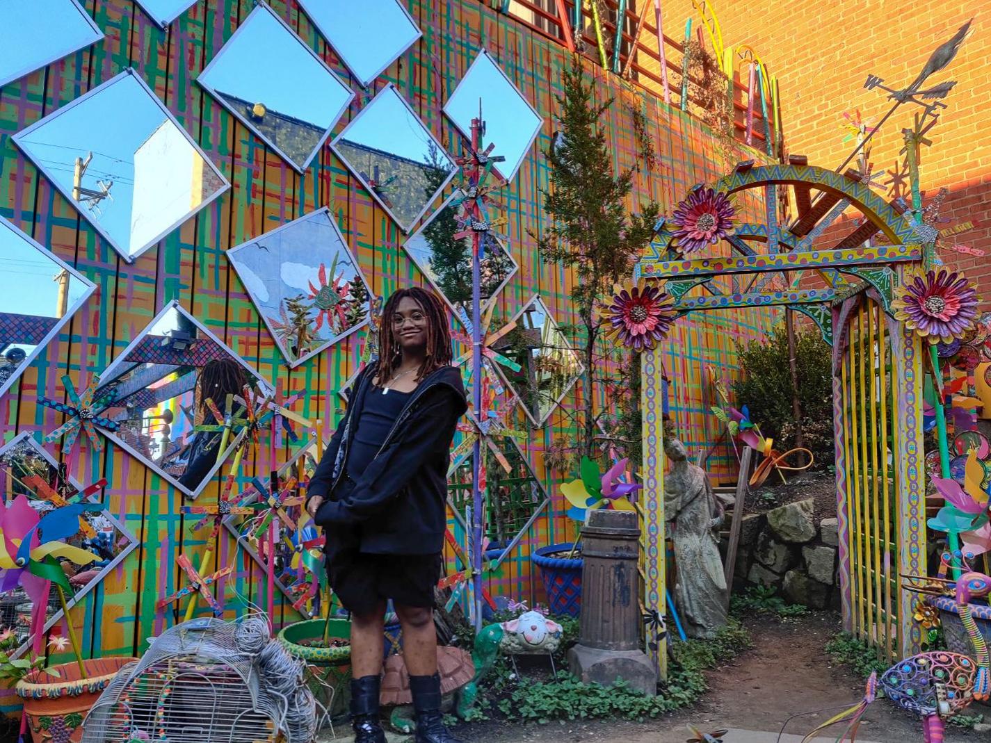 Zach Hamer-Lang, wearing a black jacket and black shorts, standing in front of a colorful wall of mirrors and art.