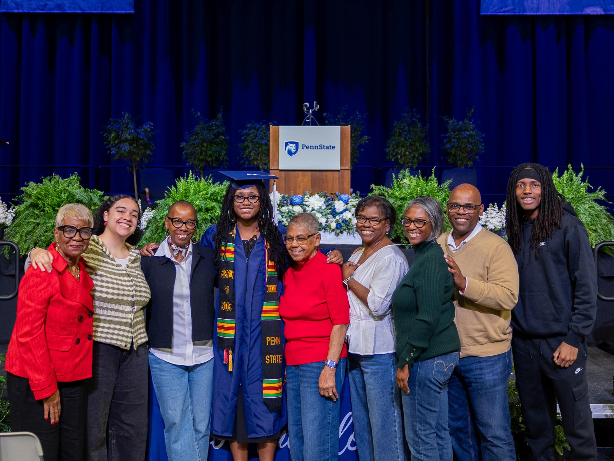 Zach Hamer-Lang, wearing a cap and gown, poses with members of his extended family in front of the Commencement stage.