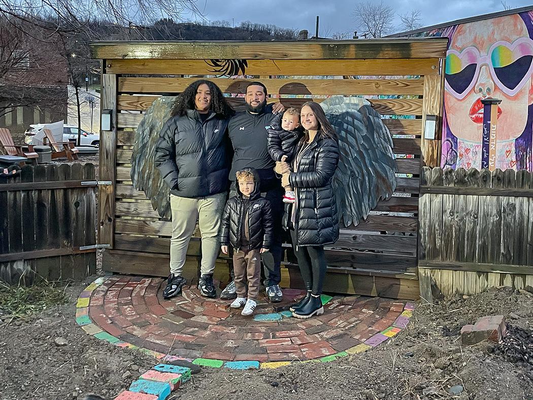 Group of adults and children standing together in front of a wooden fence with large metal wing sculptures, posed on a small circular brick area in an outdoor garden at dusk.
