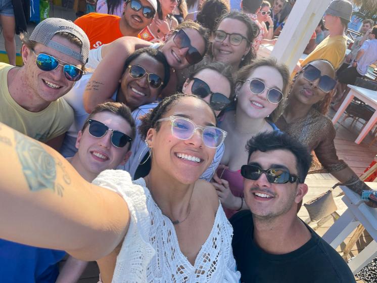 Christina  Luisa Hughes, in front, holding the camera, with a group of people gathered outdoors smile for a selfie.