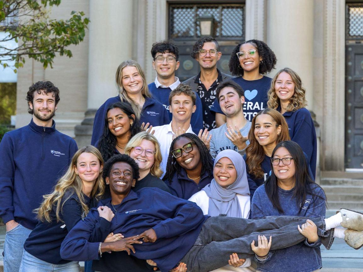 Christina Luisa Hughes and a group of fifteen young adults pose in front of a large building with columns; several people hold one person horizontally in a playful pose.