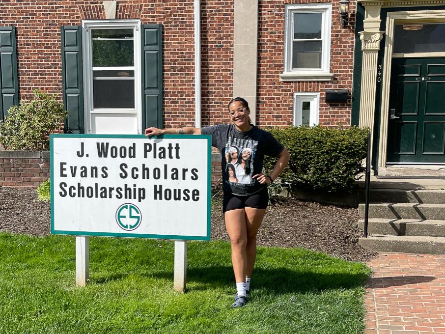 Christina Luisa Hughes stands on grass beside a white sign reading “J. Wood Platt Evans Scholars Scholarship House,” with a brick building and green shutters behind her.