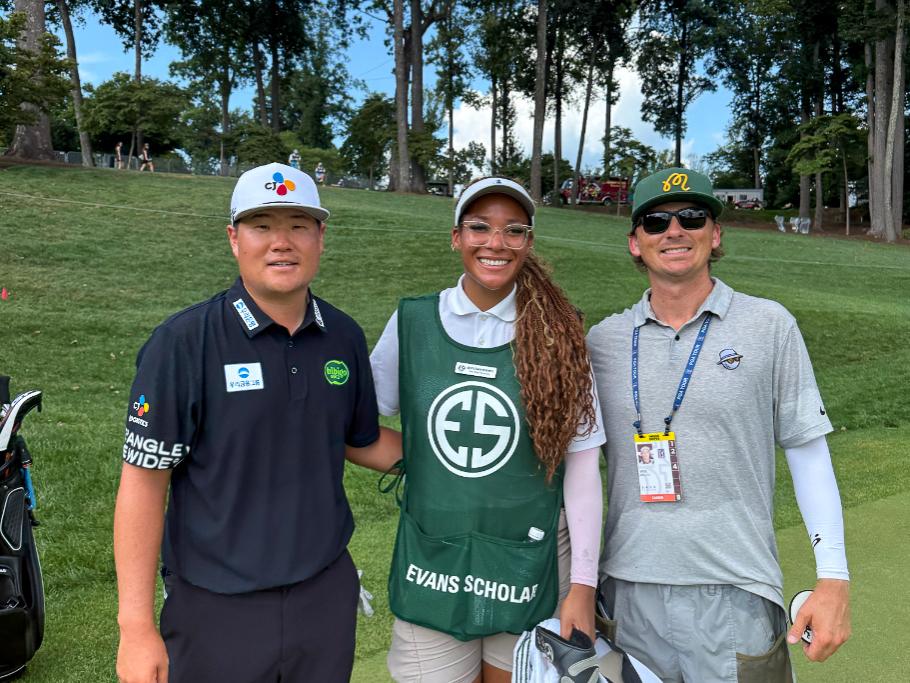Three people stand on a golf course surrounded by tall trees: a golfer in black attire, a caddie in a green “Evans Scholar” bib, and another person in gray clothing with sunglasses and event credentials.