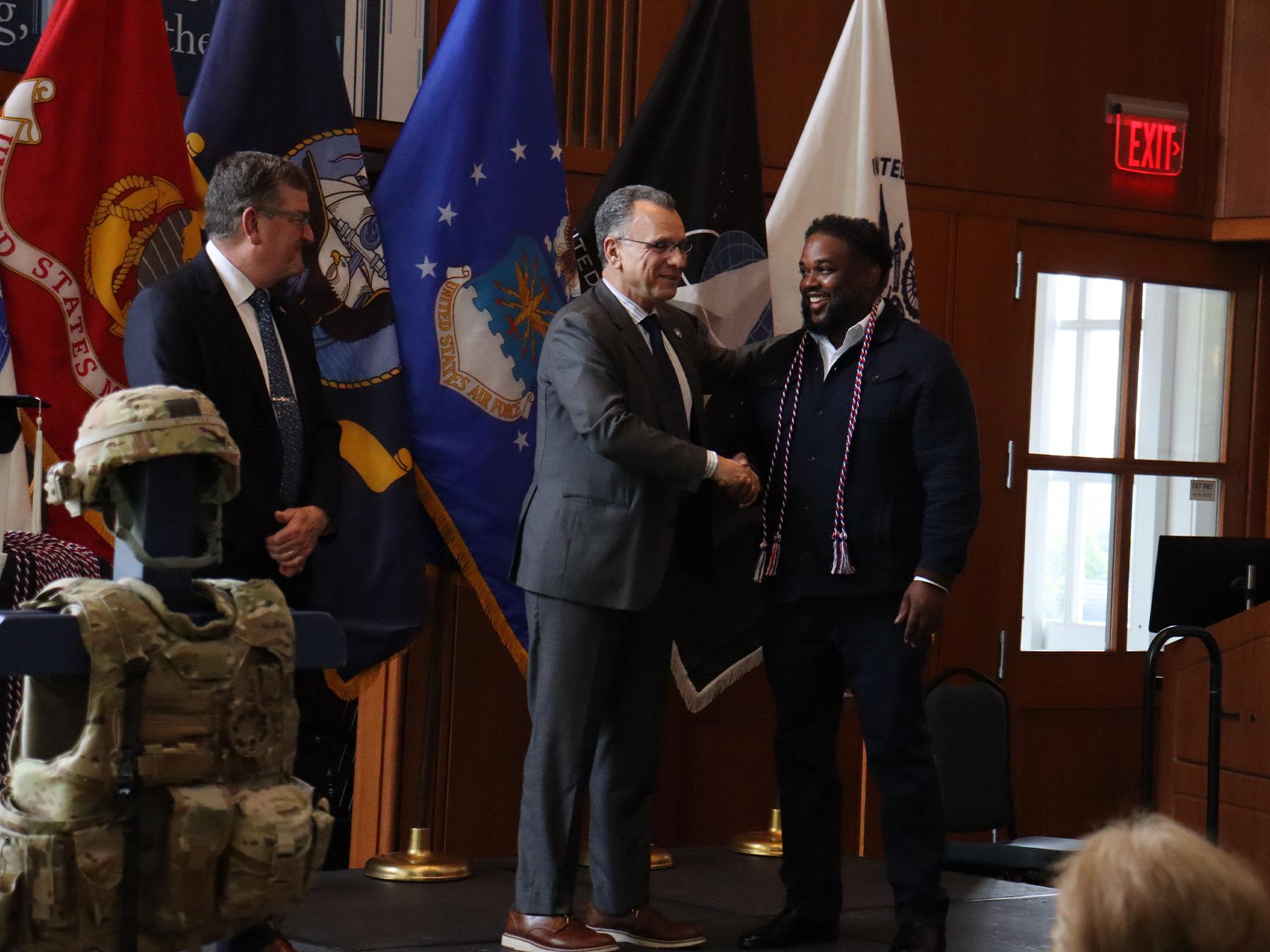 Two men shake hands in front of U.S. military flags.