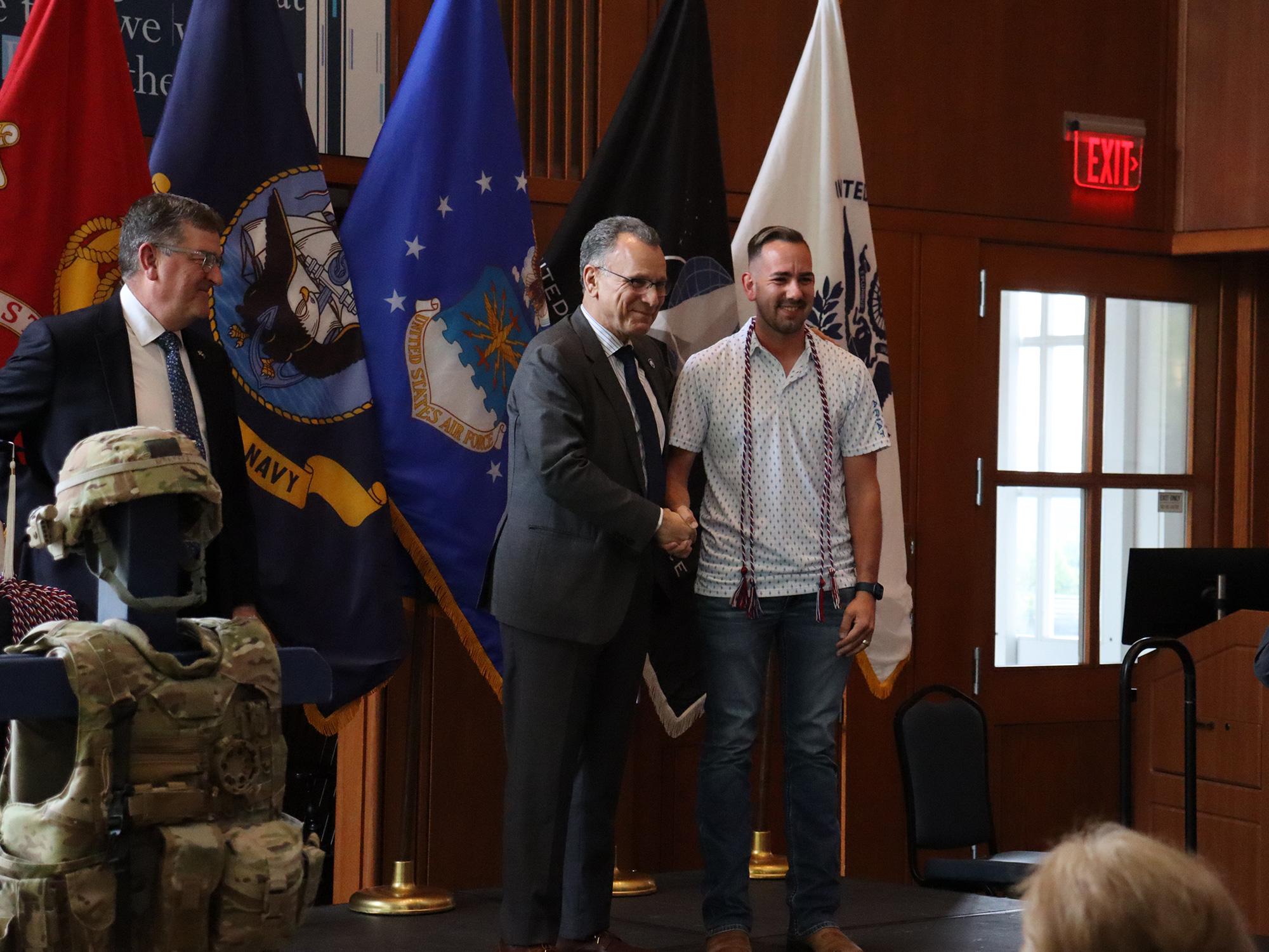 Two men shake hands in front of U.S. military flags.