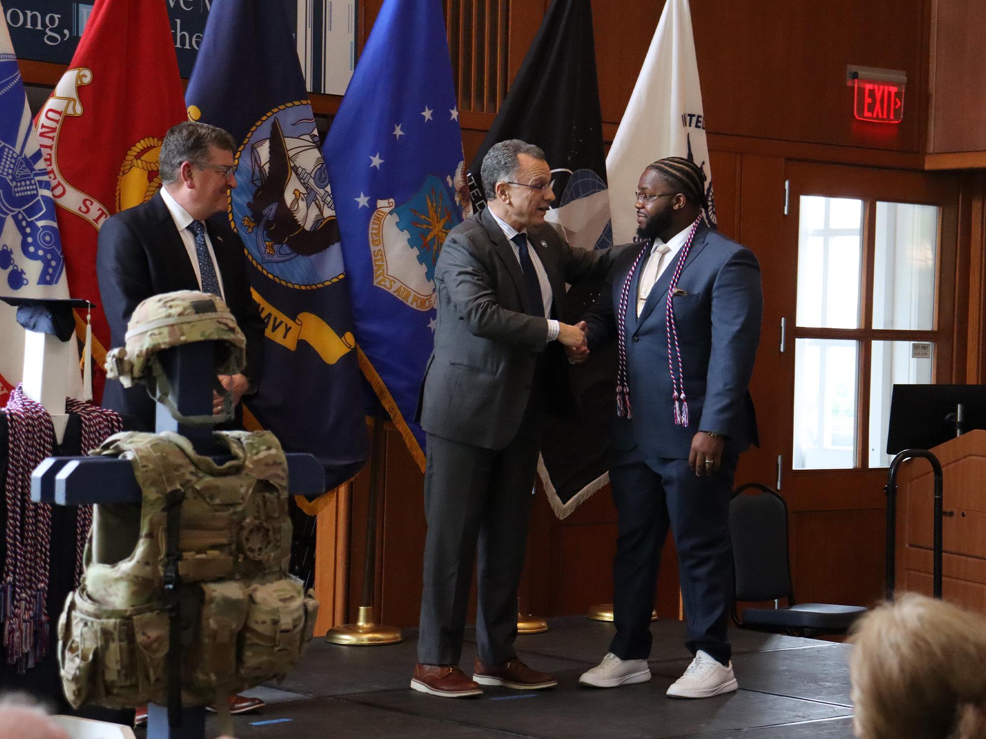 Two men shake hands in front of U.S. military flags.