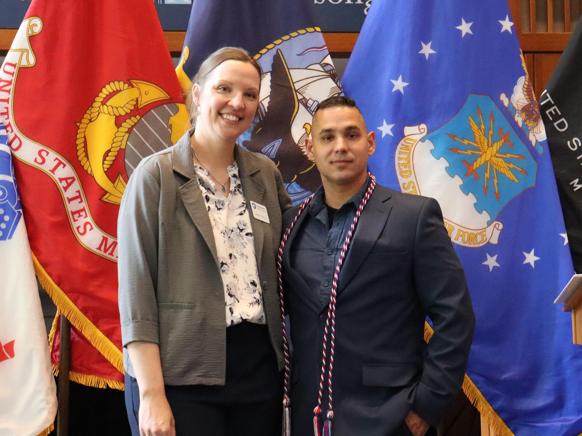 Two people pose in front of U.S. military flags.
