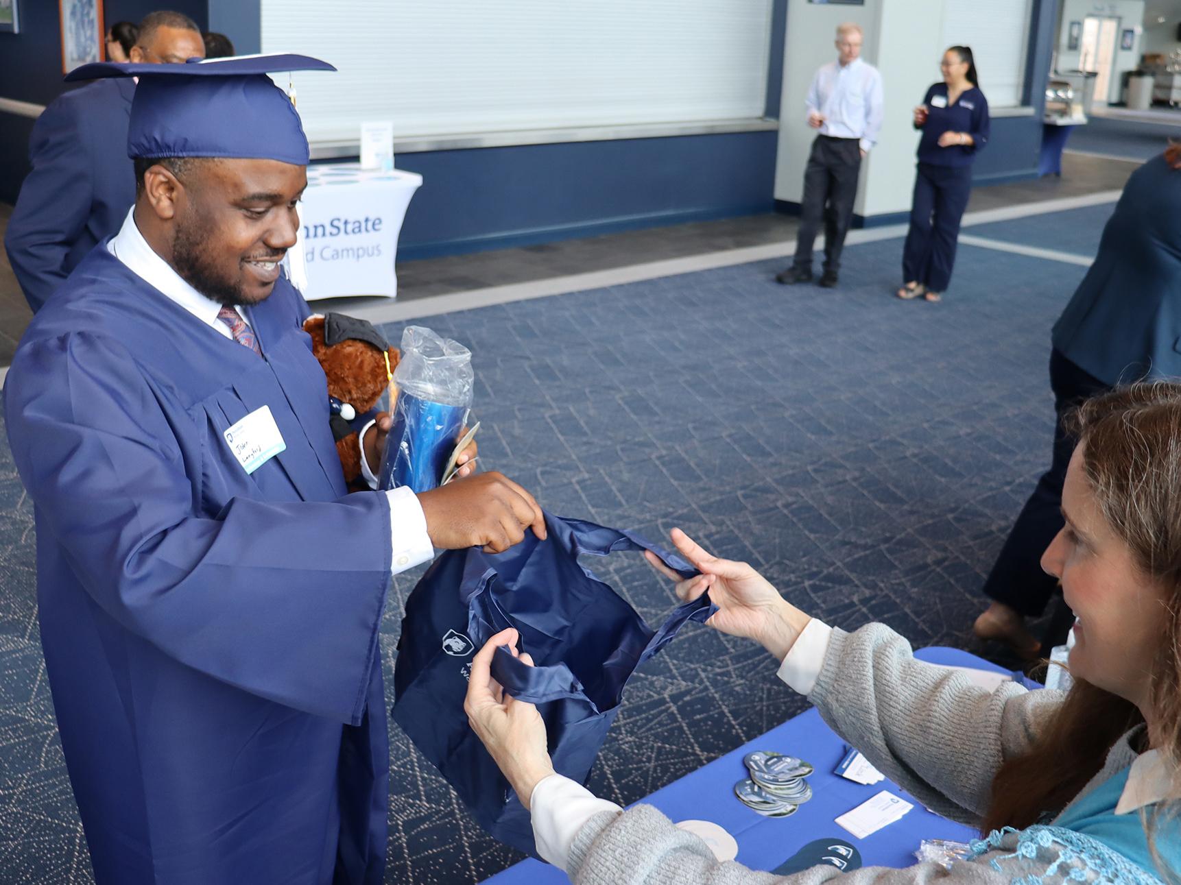 A man wearing a blue graduation cap and gown receives gifts from a woman at a table.