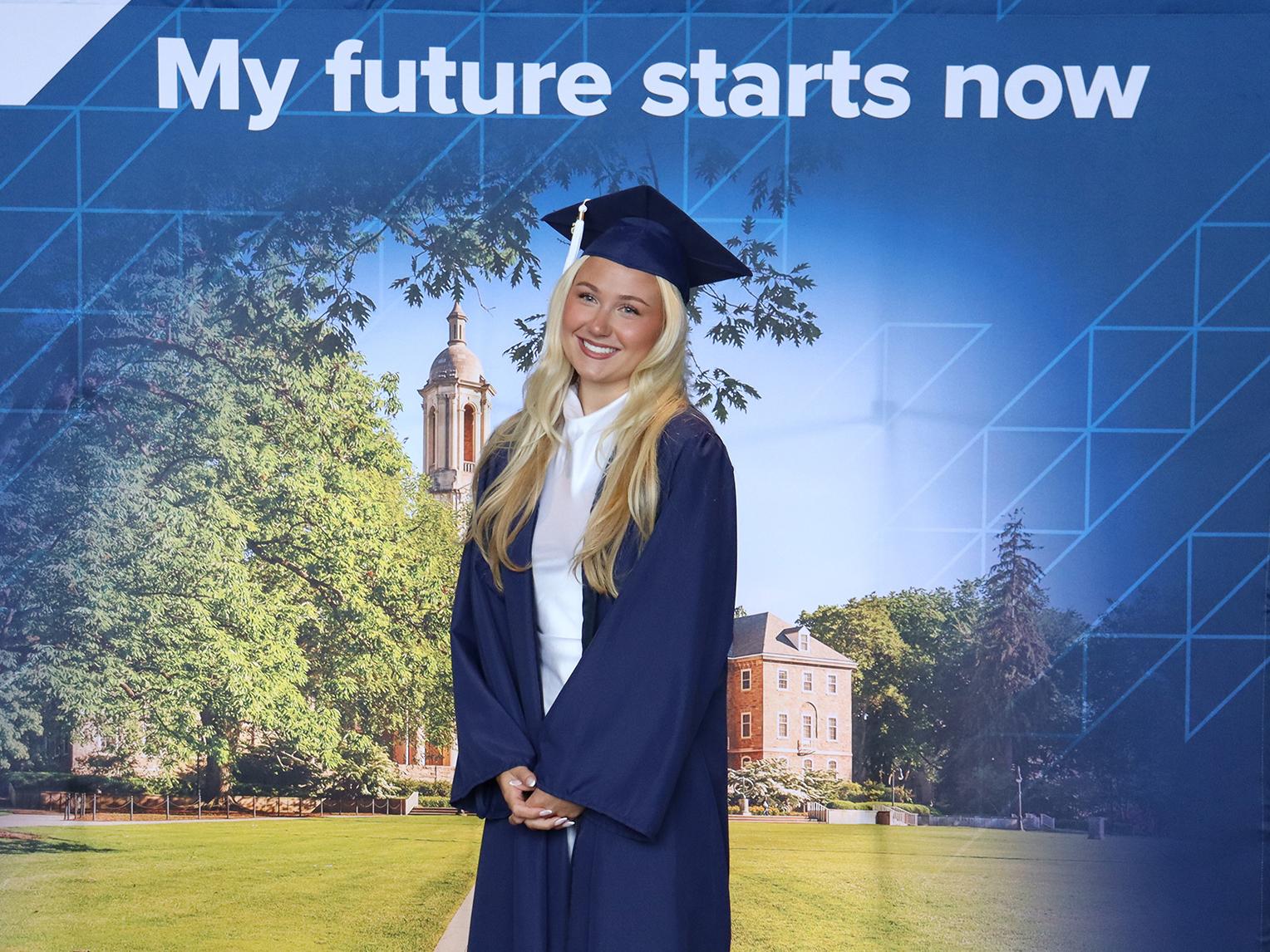 A woman wearing a blue graduation cap and gown standing in front of a Penn State photo background.