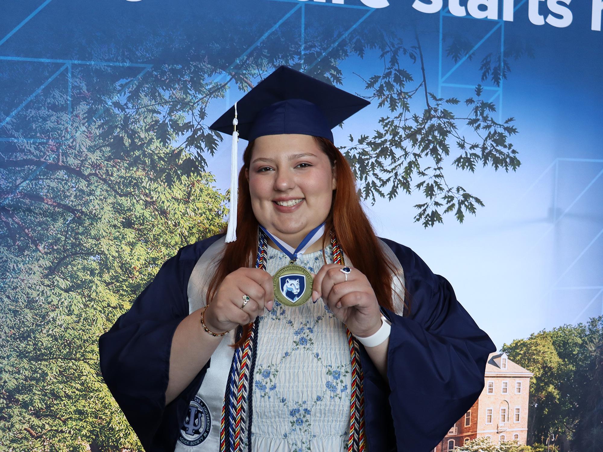 A woman wearing a blue graduation cap and gown standing in front of a Penn State photo background.