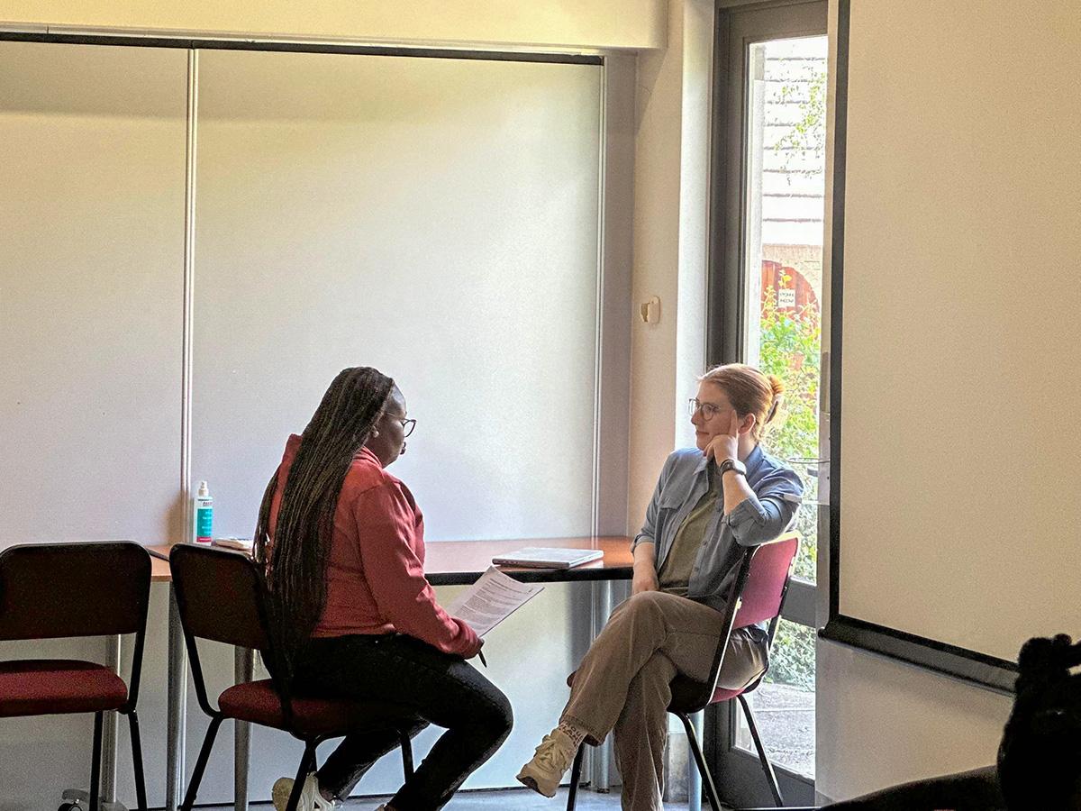 Two people sit across from each other in a room, engaged in conversation. One holds a paper, the other listens attentively. Light streams in from a window.