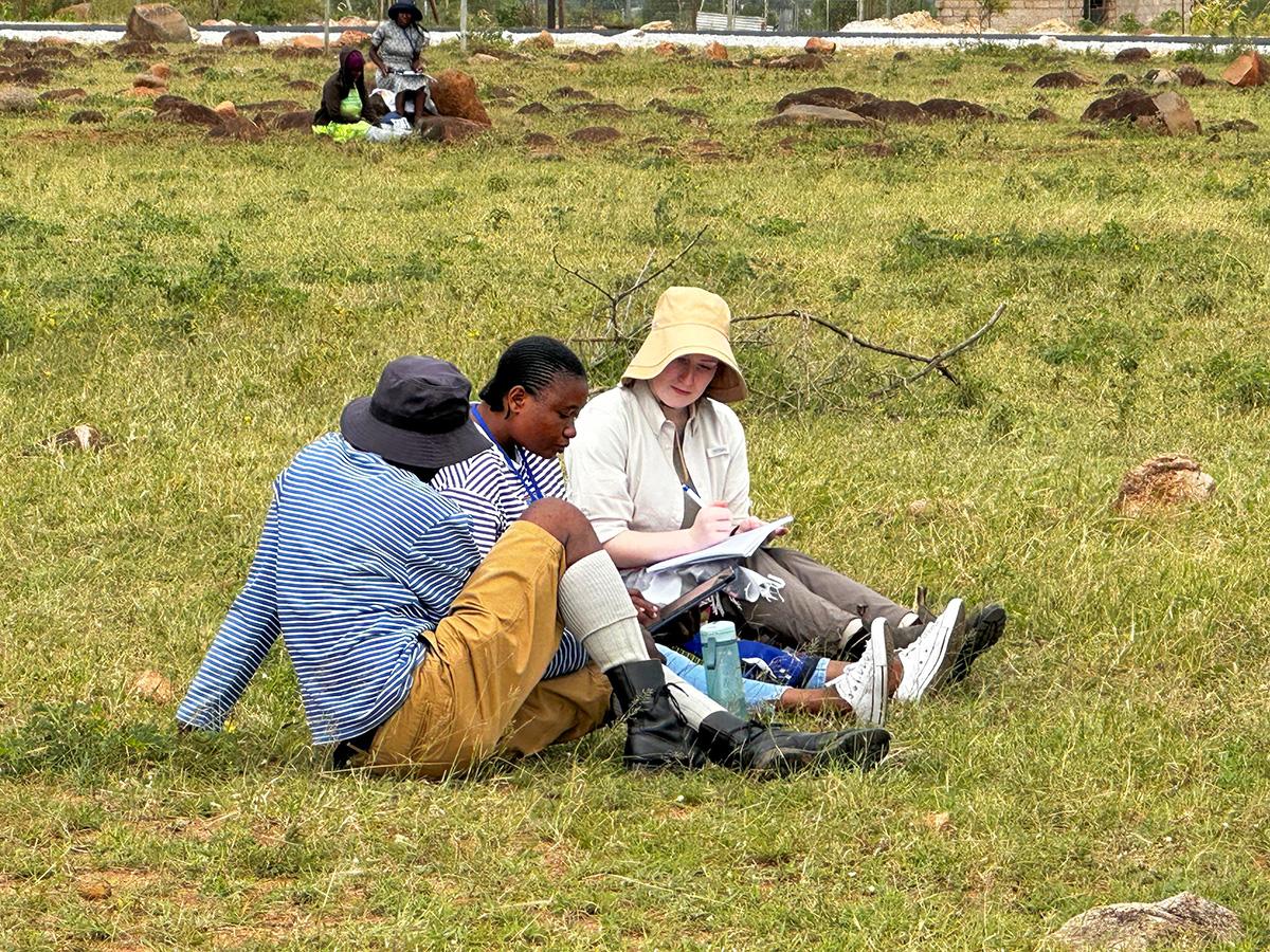 Three people sitting on grass in a field, engaged in discussion over notes. They wear casual outdoor clothing. The background shows scattered rocks.