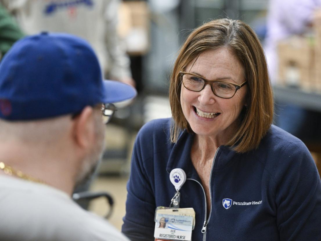 A nurse smiles at a man who is seen from behind wearing a baseball cap and glasses. The nurse has a jacket with a Penn State Health logo on it and is wearing a name badge and glasses.