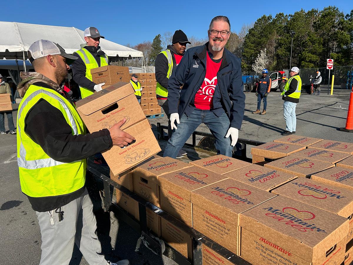 Volunteers in safety vests and gloves unload food boxes from a trailer at an outdoor donation event.