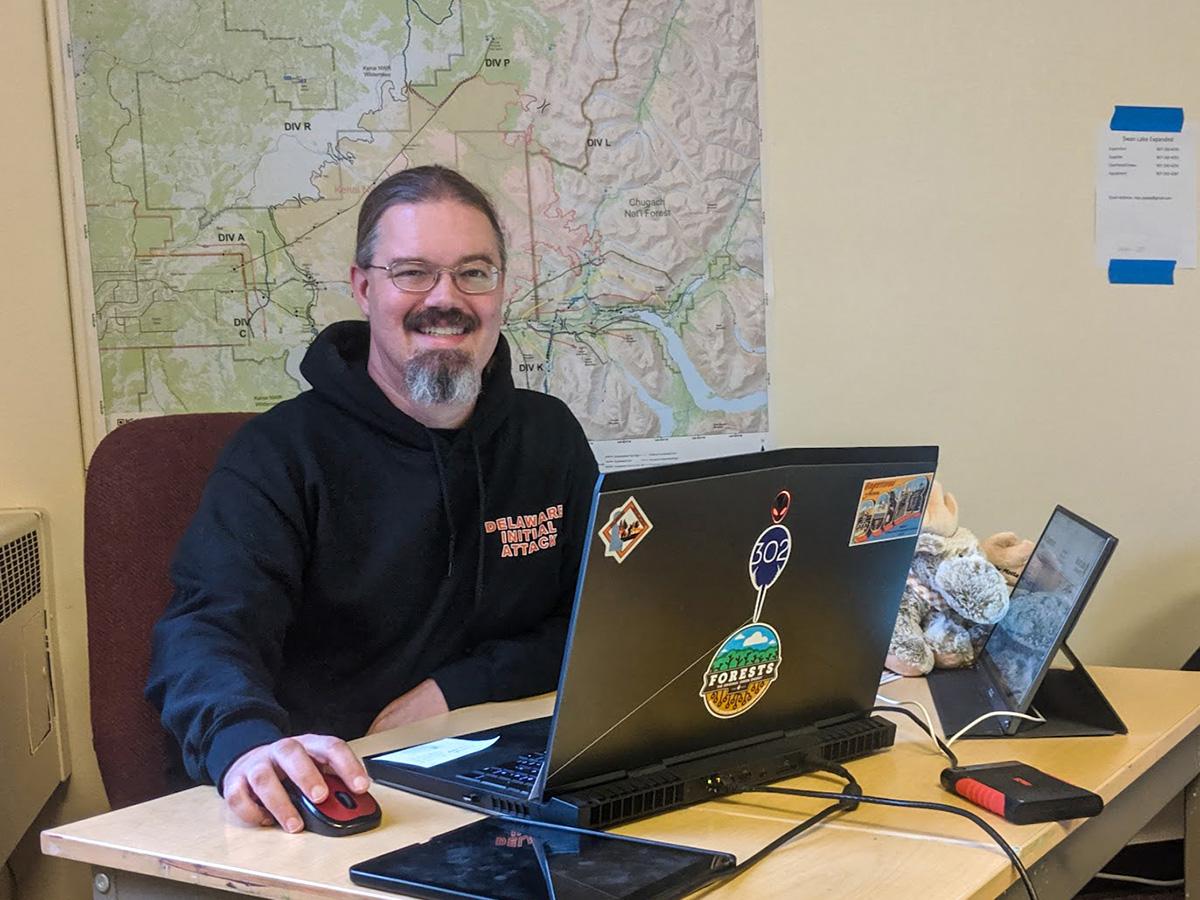 Jimmy Kroon is a smiling man with glasses and a beard sits at a desk with a laptop covered in stickers, a map on the wall behind him.