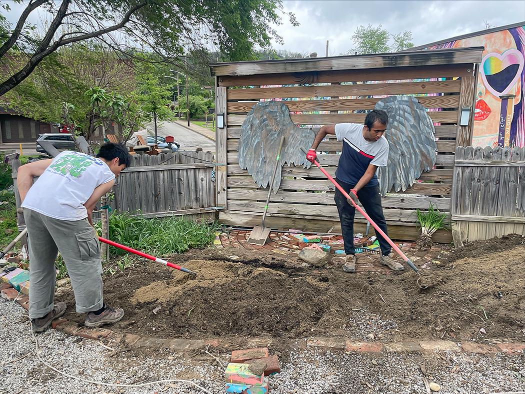 Two people raking and leveling soil in a garden bed outdoors, with large wing‑shaped artwork mounted on a wooden fence behind them.