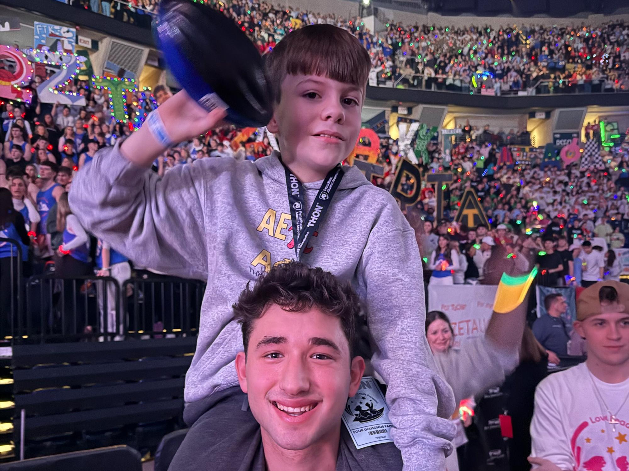 Max stands at THON on the floor wearing his fraternity sweatshirt holding a boy on his shoulders.