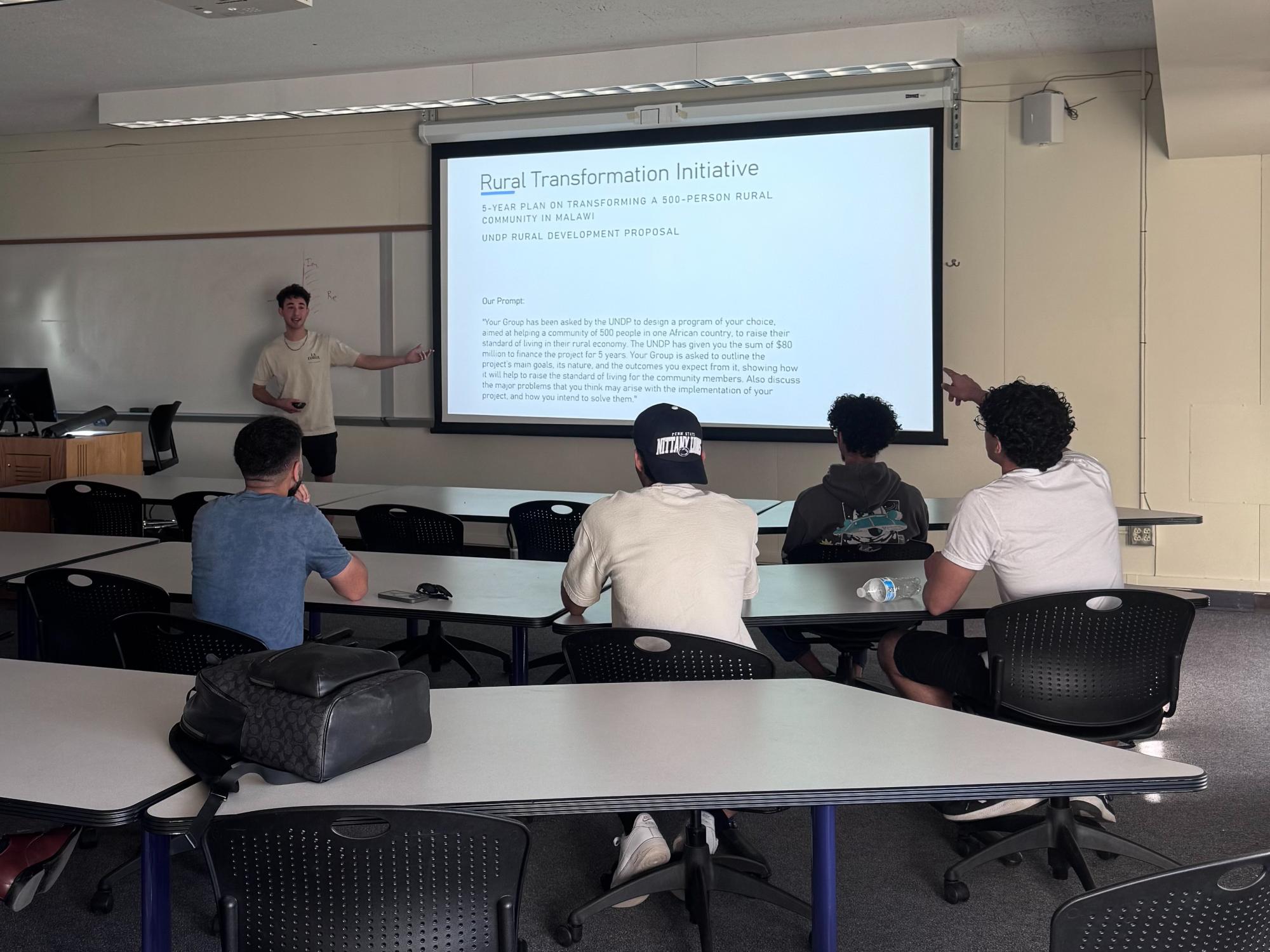 Max stands in front of a room of students pointing at the board and teaching a lesson to an African history class.