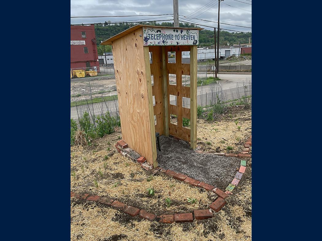 Small wooden booth labeled “Telephone to Heaven” standing outdoors, bordered by bricks and fencing, with an industrial area in the background.