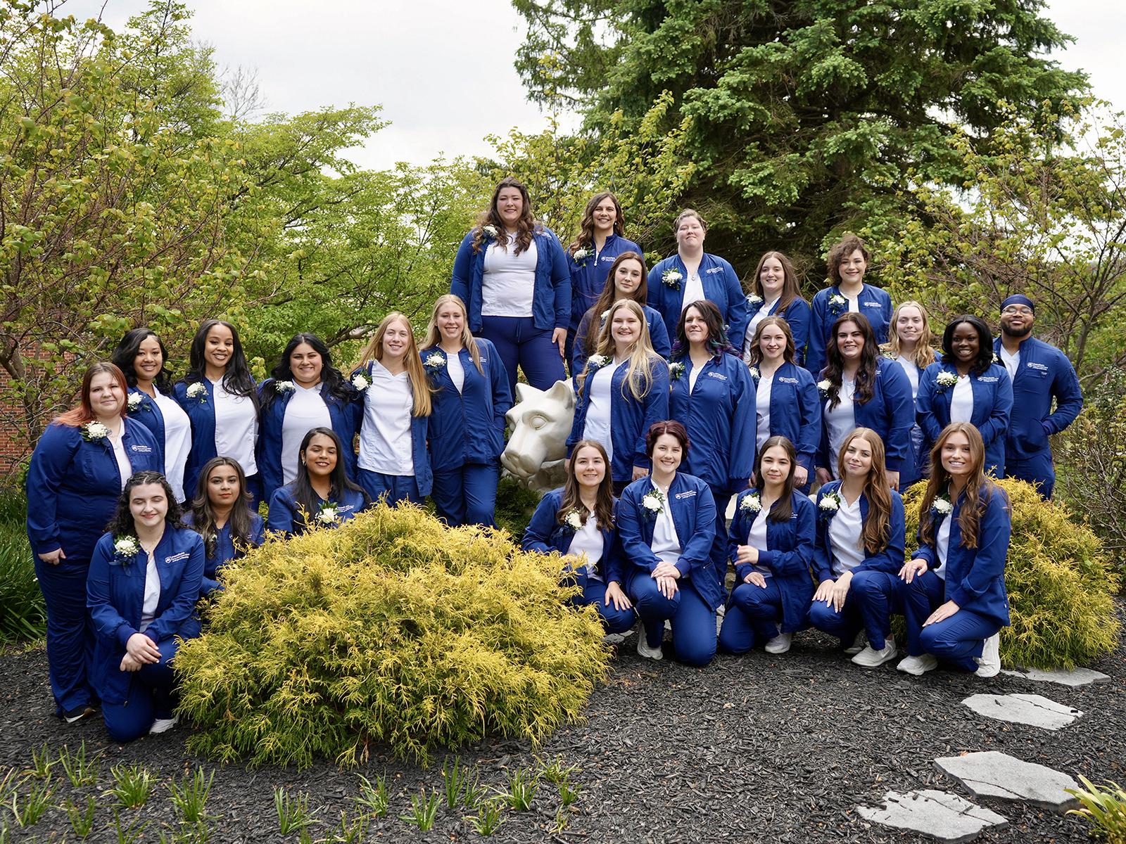 A big group of nursing students in blue robes pose for a photo outdoors on a nice day