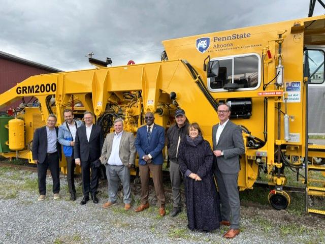 Group of people standing in front of bright yellow Penn State Altoona GRM2000 rail maintenance machine outdoors.