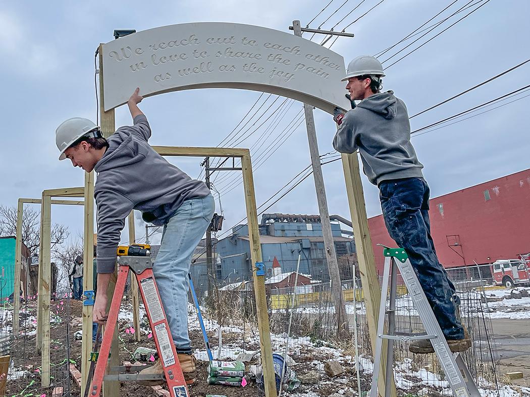 Two people wearing hard hats stand on ladders installing a curved sign on a wooden frame outdoors. The sign reads, “We reach out to each other in love to share the pain as well as the joy.” Power lines, buildings, and a winter landscape are visible in the background.
