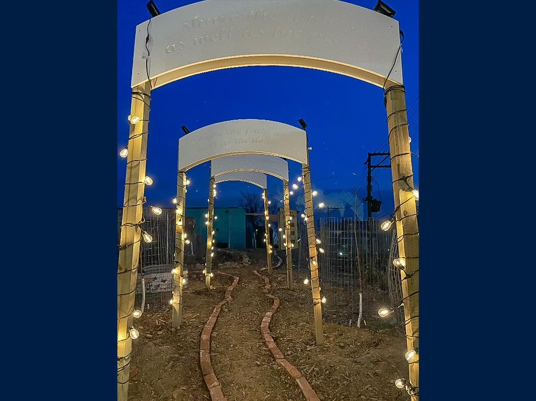 Nighttime pathway framed by wooden archways wrapped with string lights, leading along a narrow brick path.
