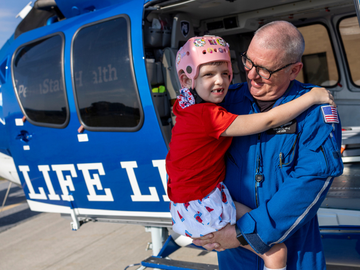 A man in a blue flight jumpsuit holds a young girl who’s wearing a helmet and smiling. They are directly in front of a Life Lion helicopter on which the side door is open.