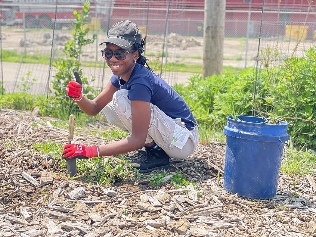 Person crouching in a garden bed, using a hand trowel to plant while giving a thumbs‑up. A blue bucket and green plants are nearby.