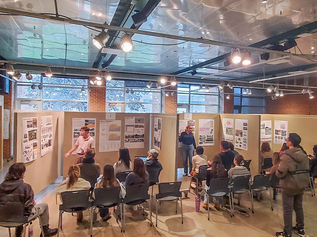 Indoor presentation with posters displayed on panels while seated attendees listen to presenters.