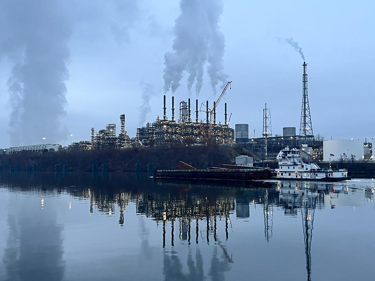 Industrial scene with a large refinery emitting smoke, reflects on a calm river. A barge and tugboat are docked in the foreground, under a cloudy sky.