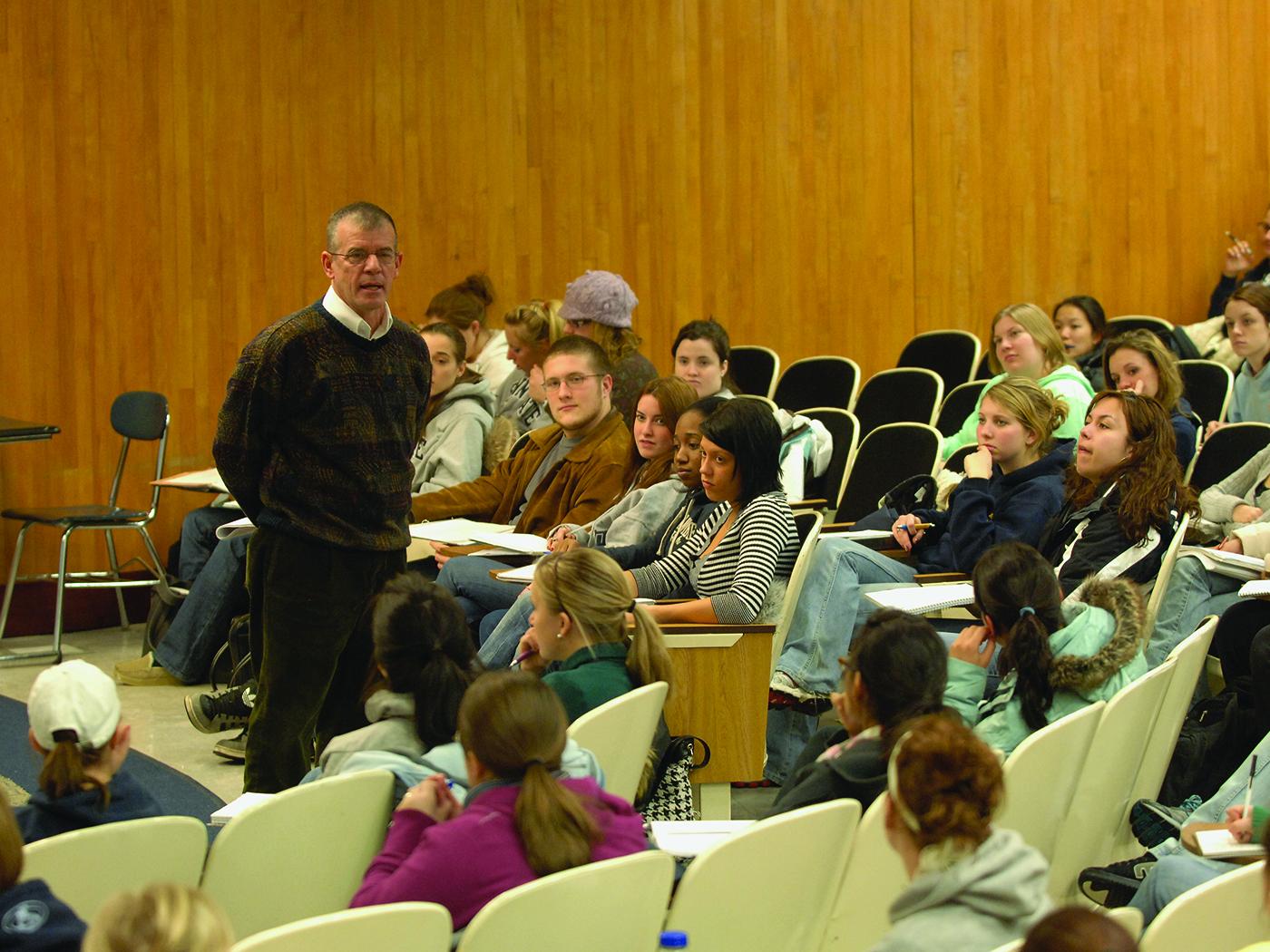 Dark haired main in a sweater talks to a large classroom of students.