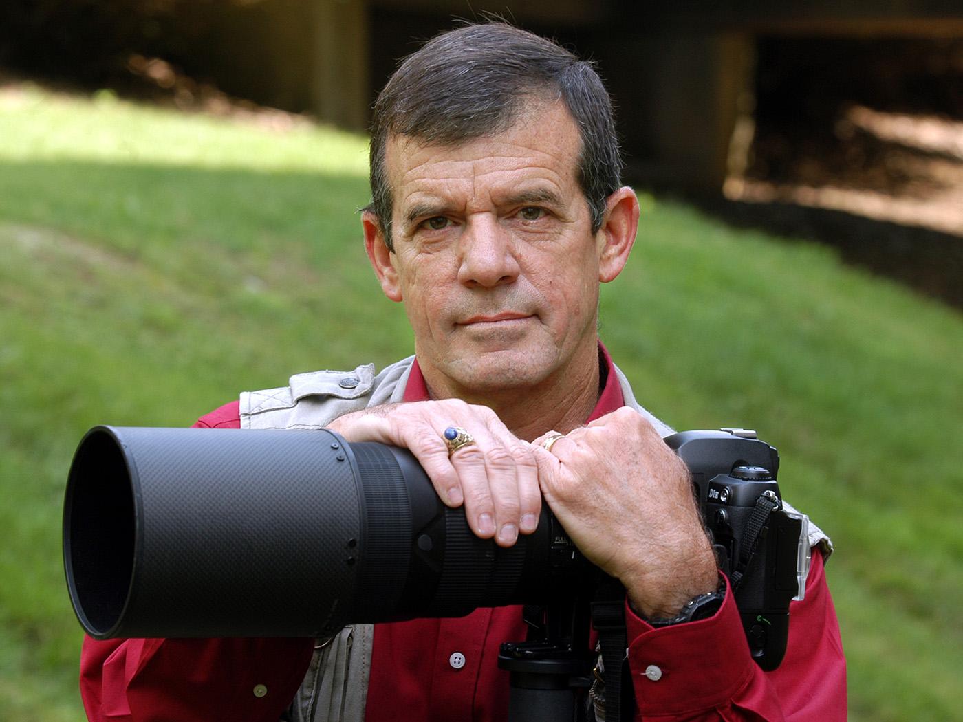 Dark haired man with red shirt and tan vest holding a camera.