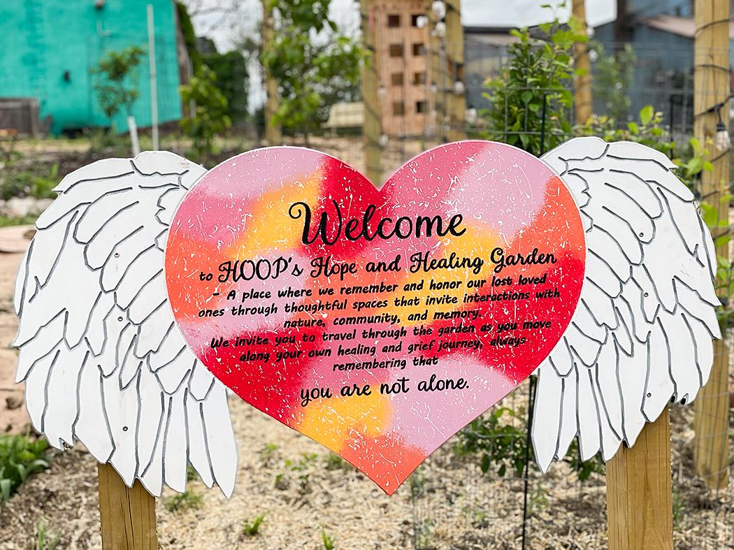Colorful heart‑shaped welcome sign with white wings mounted on wooden posts in a garden. The sign reads, “Welcome to HOOD’s Hope and Healing Garden… you are not alone,” and is surrounded by plants and soil.