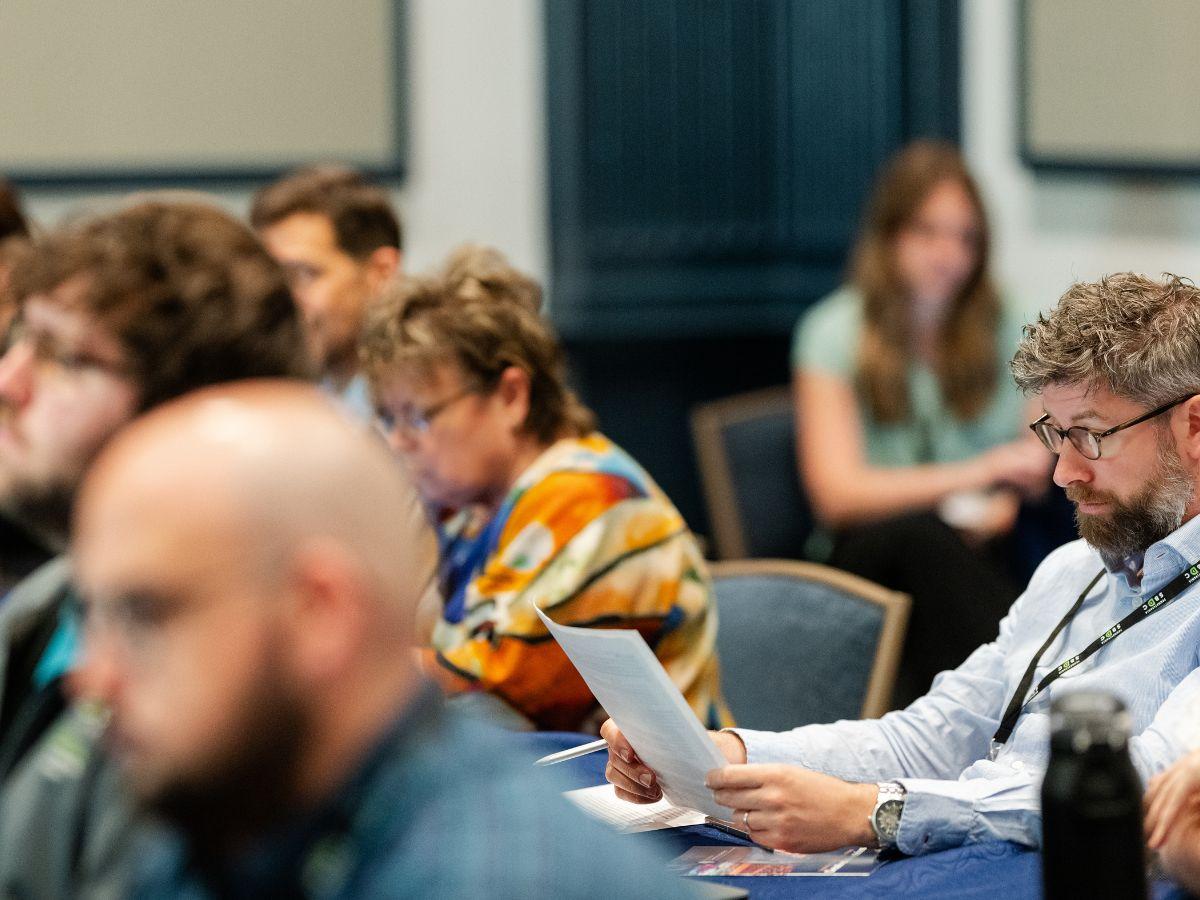 Attendees sit at tables during a conference session, reviewing materials and listening to a presentation.
