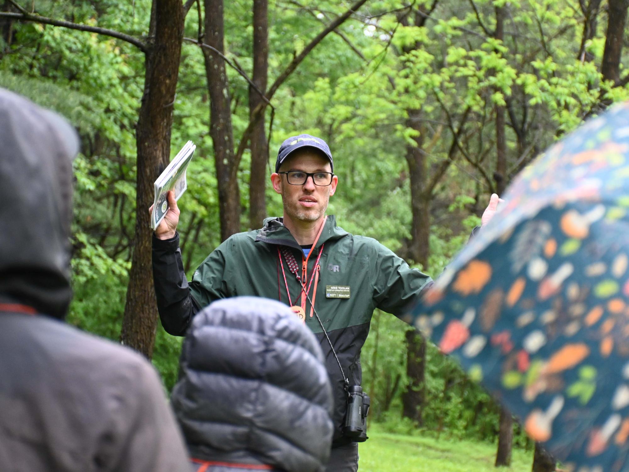 An environmental educator stands in a wooded area addressing a group, holding a booklet while speaking to attendees.