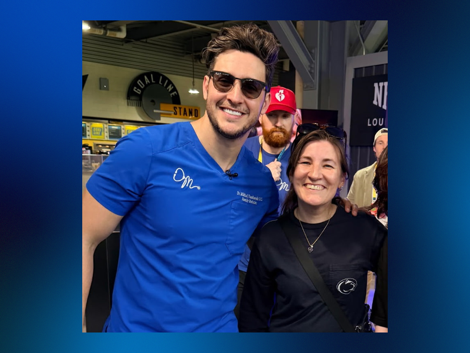A man wearing blue scrubs stands next to a woman wearing a black Penn State shirt in a stadium.