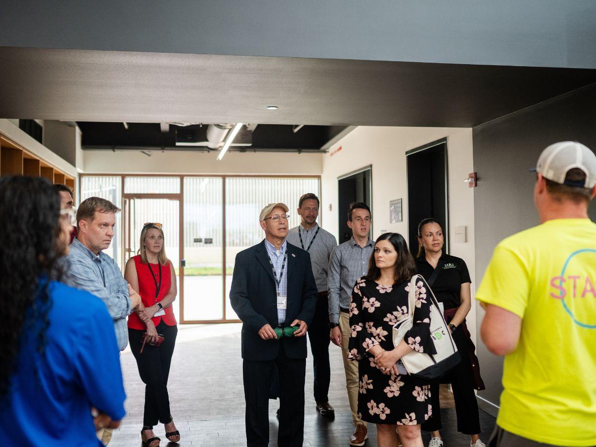 A group of professionals listens to a facility tour guide during a site visit, standing in a hallway.