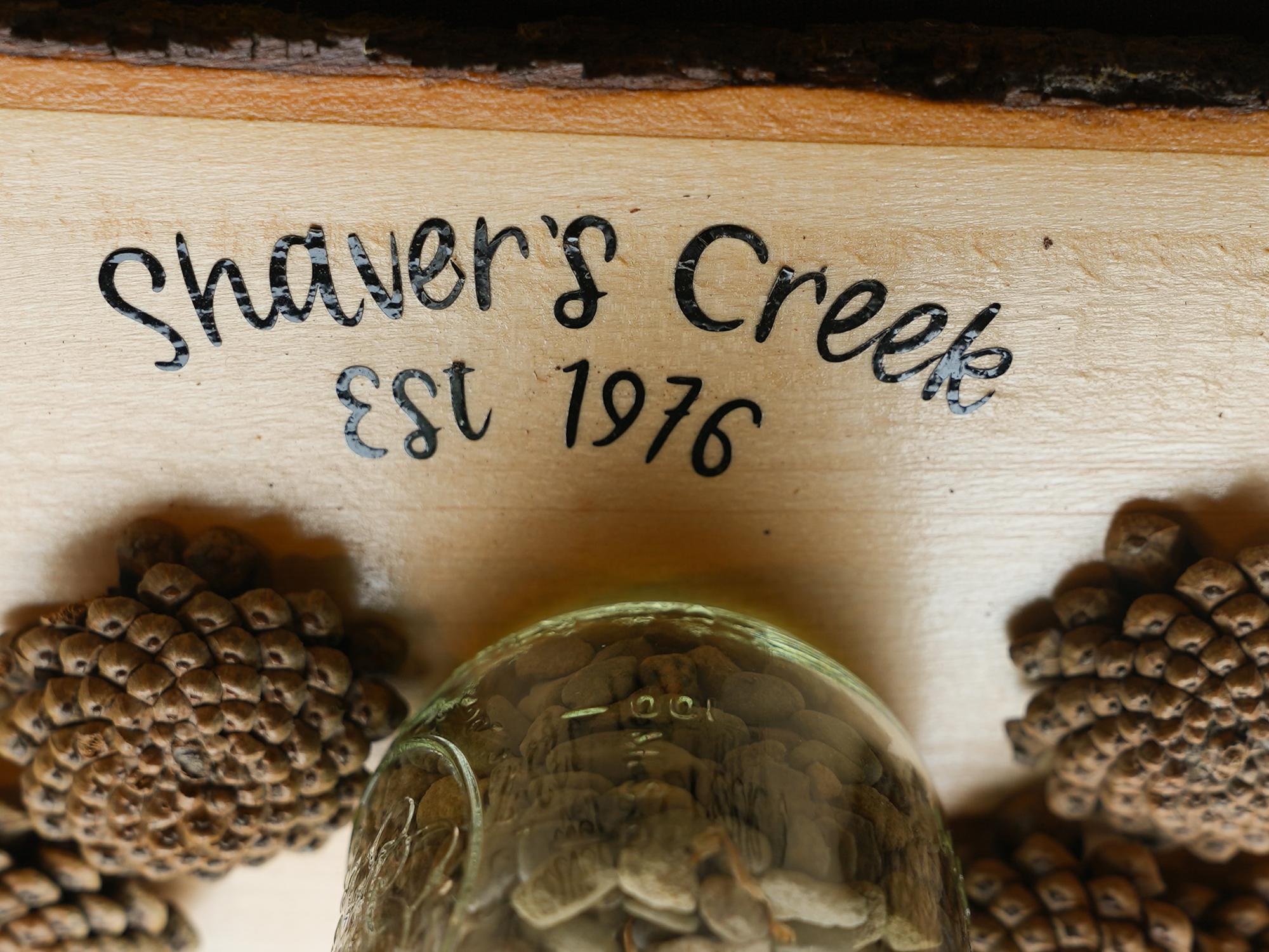 A close-up of the words “Shaver’s Creek est. 1976” engraved on wood with small natural objects arranged nearby.