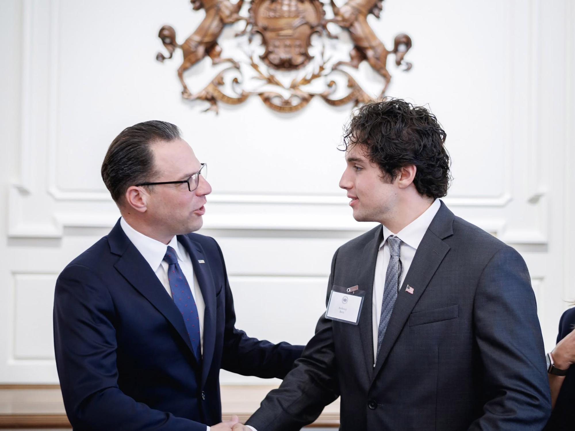 Two individuals in business attire shake hands while facing each other in a formal room. One individual wears glasses and a dark suit, and the other wears a gray suit with a name badge and an American flag pin. A decorative state crest is mounted on the wall behind them.