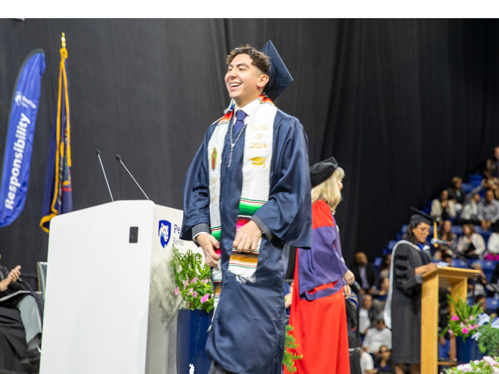 A student wearing cap and gown smiles as he receives his degree on a stage