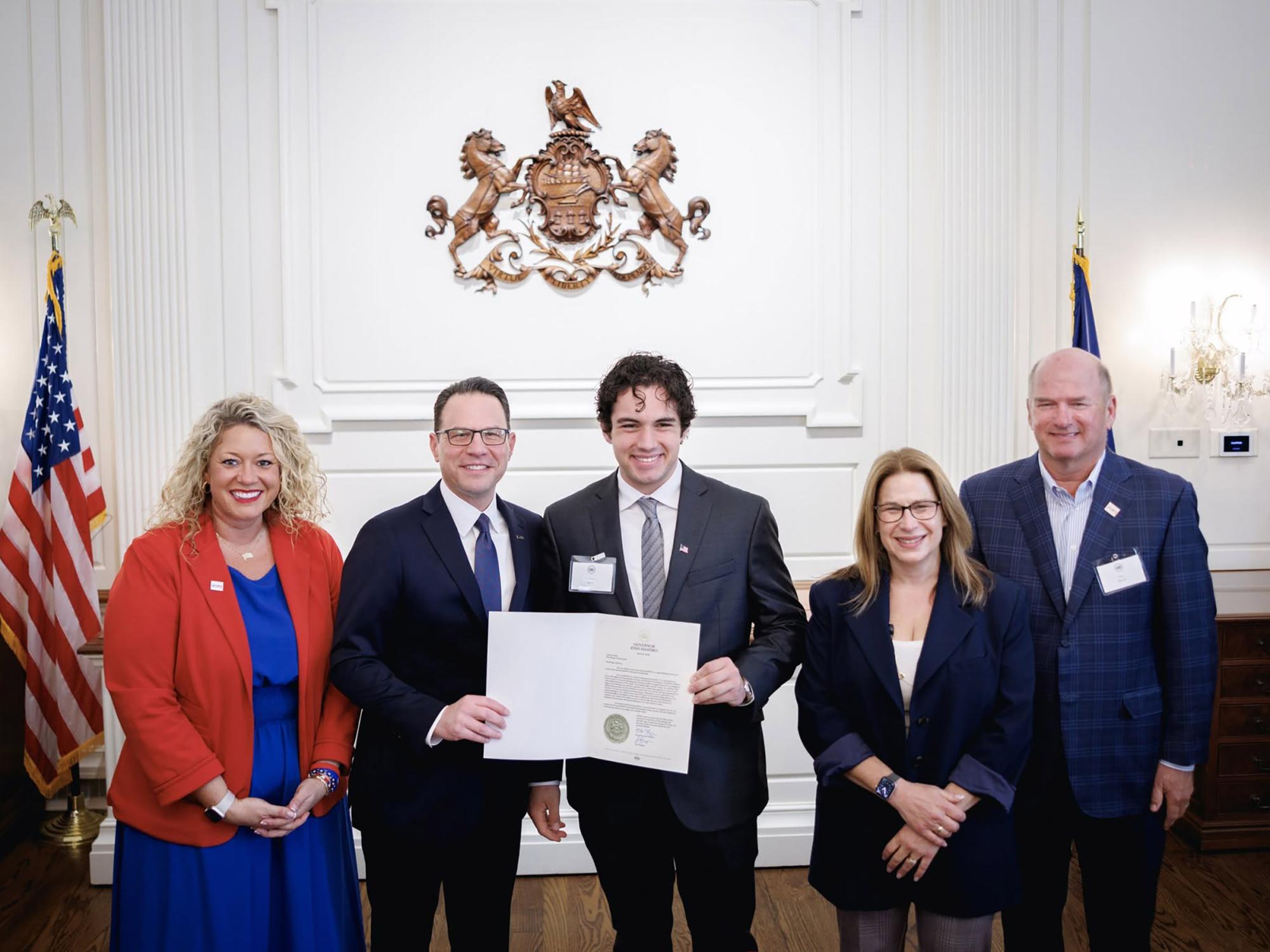 Five individuals stand in a formal room with white walls and a state crest displayed above them. The central person holds an official document while standing between two individuals in suits. Two additional individuals stand on either side. An American flag is visible to the left, and all are facing the camera.