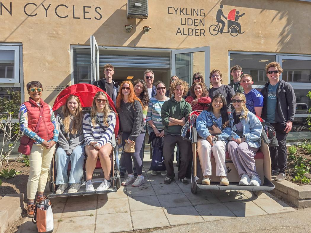 Group of approximately 18 people gathered outside a cycling organization building, posing with two bicycle rickshaws. The building behind them displays the text “CYKLING UDEN ALDER” and an illustration of a cyclist carrying a passenger.