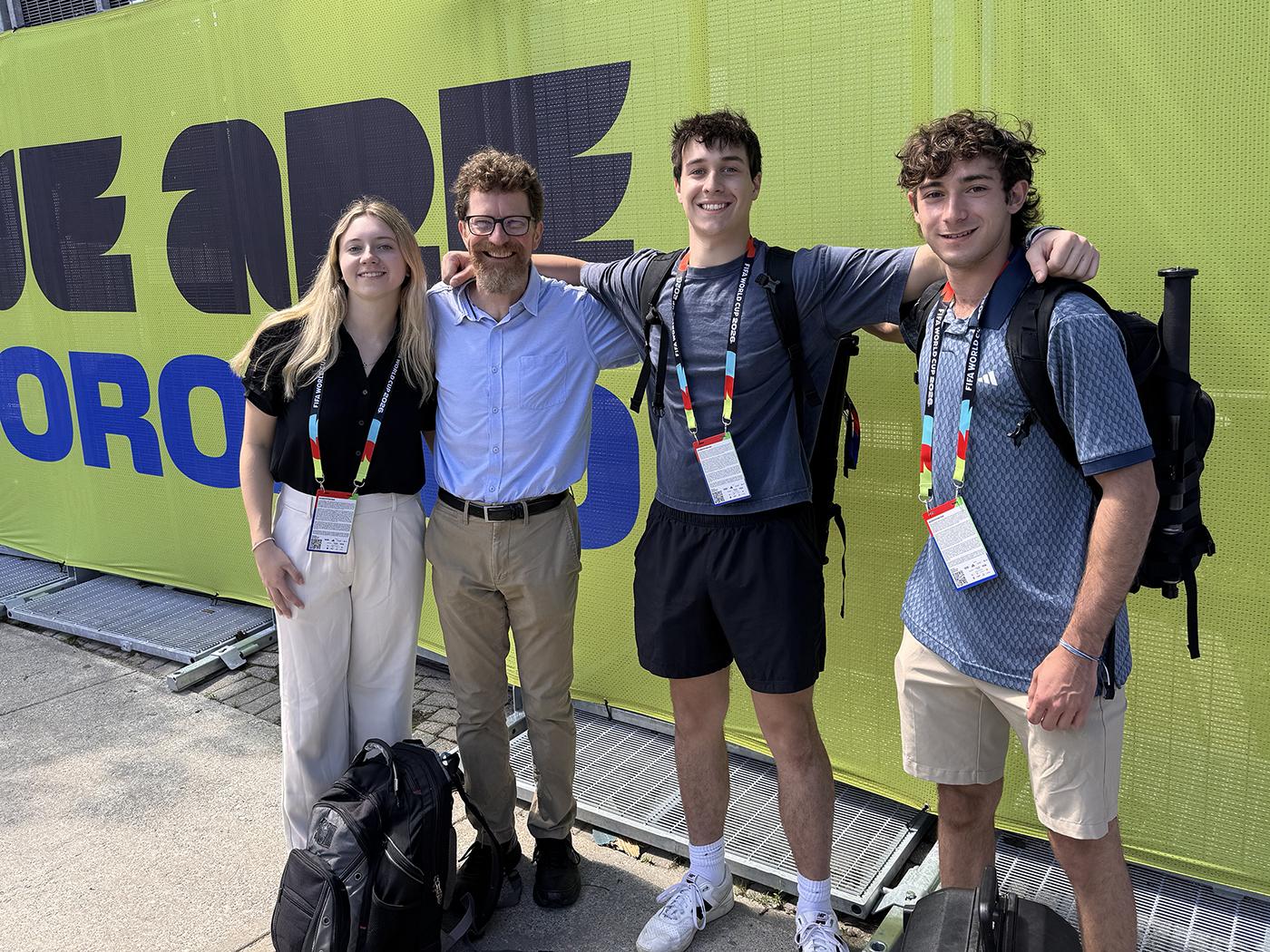 Four people standing arm in arm in front of a green sign.